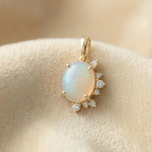 May include: A gold pendant featuring an oval opal gemstone with a halo of small, sparkling diamonds. The pendant is set against a soft, beige fabric background, creating a luxurious aesthetic.