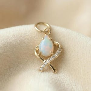 May include: A gold pendant featuring a pear-shaped opal gemstone. The pendant is set in a gold frame with a ribbon design, embellished with small, sparkling diamonds. The jewelry is displayed on a soft, cream-colored fabric.