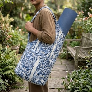 May include: A blue and white patterned yoga mat bag with a floral print, featuring a wide shoulder strap and a cylindrical shape. The bag is constructed from quilted fabric and contains a rolled blue yoga mat inside, visible at the top opening.