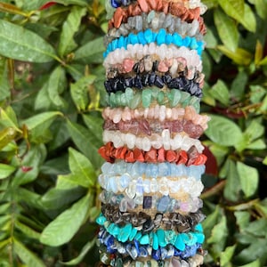 May include: A stack of gemstone chip bracelets in various colors, including turquoise, brown, and clear. The bracelets are made of small, irregular-shaped stones strung together. The background is blurred green foliage.