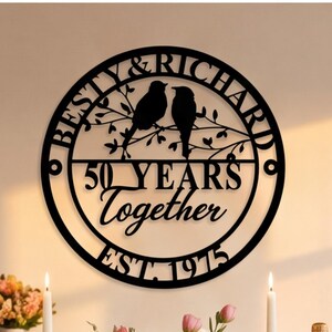 Puede incluir: Un letrero de pared circular de metal negro con el texto BESTY & RICHARD, 50 YEARS Together y EST. 1975. El diseño incluye la silueta de dos pájaros posados en una rama dentro del círculo, creando una pieza decorativa de aniversario.