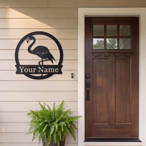 Pode incluir: Uma placa de parede circular de metal preto com a silhueta de um flamingo e uma faixa horizontal no centro com o texto Your Name em uma fonte serifada branca, montada em uma parede externa clara ao lado de uma porta de madeira escura.