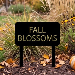 Op de afbeelding: Een rechthoekig zwart tuinbord met afgeronde hoeken voorzien van de tekst FALL BLOSSOMS in een goudkleurig schreefloos lettertype. Het bord wordt ondersteund door twee zwarte palen in de grond, geplaatst voor siergrassen.