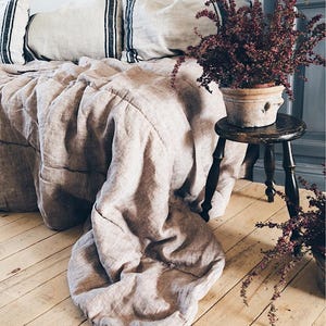 Peut inclure: Un couvre-lit beige et des oreillers à rayures vertes et noires. Un petit tabouret en bois supporte une plante en pot avec des feuilles bordeaux. L'ensemble est posé sur un plancher en bois clair.
