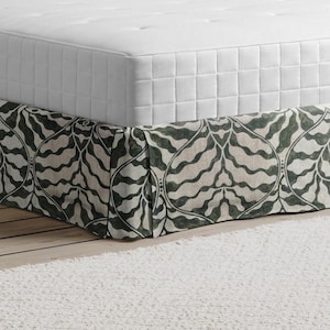 Può includere: Un letto con un materasso bianco e una gonna da letto fantasia. La gonna da letto presenta un motivo ripetuto nei toni del verde e del bianco. Sono visibili anche un piccolo comodino in legno e una pila di libri.