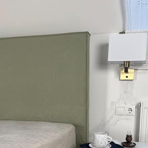 May include: A white bedside table with two drawers and brass ring handles. A patterned cup and saucer rest on a navy cloth. A bed with a green headboard and base is in the background. A white lamp is mounted on the wall.