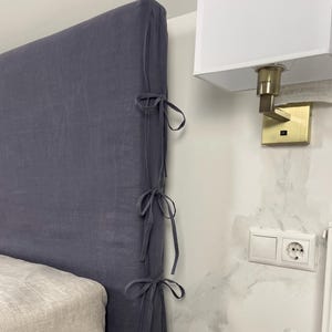 May include: A dark gray linen headboard cover with tie closures on the side. The headboard cover is on a bed with a light beige mattress. A white nightstand holds a speckled mug and saucer, a candle, and a dark blue cloth.