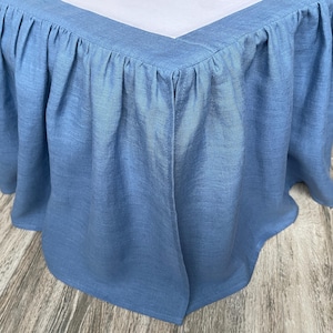 May include: A light blue bed skirt with a gathered, ruffled design. The fabric has a textured appearance and drapes around the bed frame. The bed skirt is made of a soft, lightweight material.