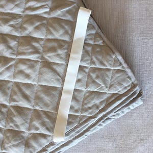 Puede incluir: Una manta acolchada de color gris claro con una cinta blanca. La manta tiene un patrón de diamantes y está doblada. La cinta está centrada en la manta y está hecha de un material liso. La manta está sobre una superficie gris claro.