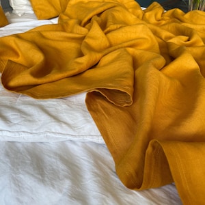 Puede incluir: Primer plano de una cama con sábanas blancas y una manta de lino amarillo mostaza. La manta está drapeada sobre la cama, creando pliegues y sombras suaves. Se ve una almohada a juego. La tela tiene textura.