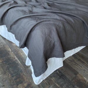 Könnte beinhalten: Eine dunkelgraue Bettdecke aus Leinen, die über einen weißen Bettrock drapiert ist. Das Bett steht auf einem dunklen Holzboden. Der Leinenstoff hat ein weiches, strukturiertes Aussehen, und der weiße Bettrock hat einen gewellten Rand.