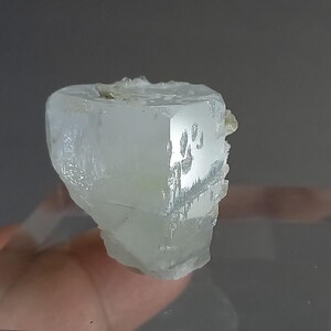 May include: A pale blue, crystalline mineral specimen with a geometric shape. The crystal has a translucent quality, with visible internal structures and a slightly rough texture. Small, light green mineral deposits are visible on the top and side.