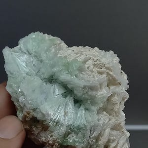 Puede incluir: Primer plano de un espécimen mineral con un grupo de cristales verde claro en forma de aguja. Los cristales se asientan sobre una matriz rugosa de color blanquecino. La textura general es áspera y cristalina.
