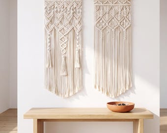 May include: Two off-white macrame wall hangings featuring intricate knotted patterns and hanging tassels, suspended from wooden dowels. The wall hangings are displayed above a light-toned wooden bench with a terracotta bowl placed on top.