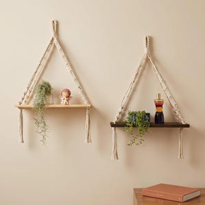 May include: Two wall-mounted shelves suspended by cream-colored macrame rope. The left shelf is light wood holding a small plant and a figurine. The right shelf is dark wood holding a green plant and a glass bottle. Both feature knotted rope.