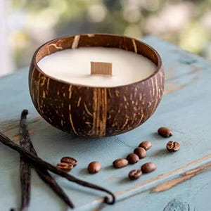 May include: A coconut shell candle with a wooden wick, filled with white wax. The candle sits on a blue surface, with coffee beans and vanilla beans scattered around it. The candle is a natural, rustic home decor item.