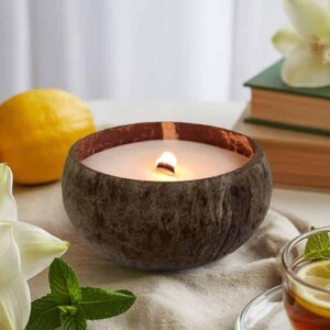 May include: A lit candle in a brown coconut shell. The candle has a white wax and a wooden wick. The shell sits on a table with lemons, flowers, and a cup of tea.