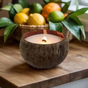 May include: A lit candle in a coconut shell sits on a wooden surface. The candle has a white wax interior and a small flame. A basket of lemons, limes, and oranges with green leaves is in the background.