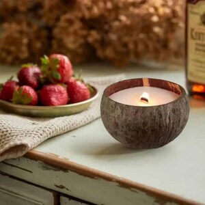 May include: A lit candle in a coconut shell sits on a distressed white table. A small plate of fresh strawberries and a bottle of Kentucky bourbon are in the background. The candle has a white wax and a wooden wick.