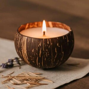 May include: A lit candle in a halved coconut shell. The candle has a white wax and a flickering flame. The coconut shell is brown with a textured surface. Lavender sprigs and dried plant material are on the linen cloth.