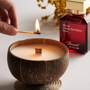 May include: A lit match ignites a candle in a coconut shell. The candle has a wooden wick and sits on a woven coaster. A red perfume bottle with a gold cap is in the background.
