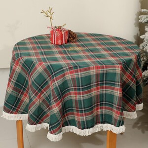 May include: A round table with a green, red, and white plaid tablecloth. The tablecloth has a white fringe trim. A small gift box and decorative sprig sit on the table. The table has four wooden legs.