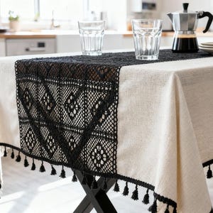 Könnte beinhalten: Cremefarbene Tischdecke mit schwarzem Spitzentischläufer mit geometrischem Muster und Quastenbesatz. Zwei Gläser, eine Kaffeemaschine und Teller stehen auf dem Tisch. Der Tisch steht auf einem schwarzen Metallgestell.