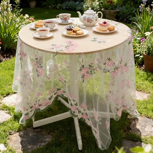 Könnte beinhalten: Ein runder Tisch für Tee in einem Garten. Der Tisch hat eine cremefarbene Tischplatte mit rosa floralen Akzenten und ist mit einer durchsichtigen, bestickten Tischdecke bedeckt. Teetassen, eine Teekanne und Gebäck sind auf dem Tisch angeordnet.