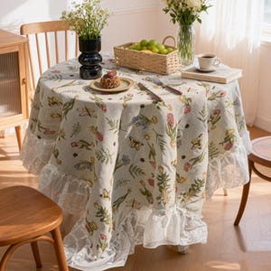 May include: A round tablecloth with a floral and butterfly design, edged with white lace. The table is set with a cake, cutlery, and a coffee cup. A vase of flowers and a fruit basket are also on the table.