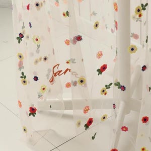 May include: Sheer white curtain with embroidered colorful flowers. The word "far" is written in red script on the curtain. The flowers are red, yellow, and purple.