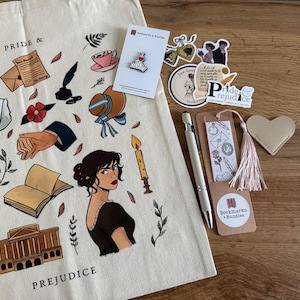May include: A cream-colored tote bag with illustrations from "Pride & Prejudice" sits next to a collection of themed items. These include stickers, a bookmark with a tassel, a pen, a heart-shaped leather item, and a pin.