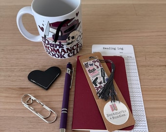 May include: A collection of book-themed items, including a white mug with "Dark Romance Book Club" text and graphics, a black heart-shaped coaster, a purple pen, a bookmark, a reading log, and a paperclip. The bookmark has a tassel and the text "Dark Romance".