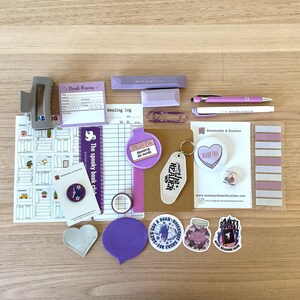 May include: A selection of purple and white stationery items, such as a pen, highlighter, paper clip, and stickers, arranged on a wooden surface. Some items feature text, including "Book Review" and "BOOK ERA".