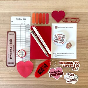 May include: A collection of stationery and accessories, including a red notebook, pens, sticky notes, and stickers. The items are arranged on a wooden surface. The stickers have phrases like "Enemies to Lovers" and "I'm here for Red Flags."