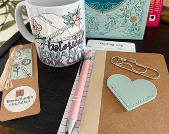 Op de afbeelding: Een witte mok met de tekst "Historical Romance Book Club" en bloemaccenten, een bladwijzer, een notitieboek, een pen, een paperclip en een hartvormige bladwijzer. Op de achtergrond staat een boek met de titel "Seven Novels". De items zijn gerangschikt op een zwart oppervlak.