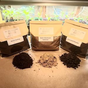 Pode incluir: Três sacos de corretivos do solo rotulados "Worm Castings", "Spent Mushroom Substrate" e "Inoculated Biochar". Cada saco é bege e preto, com uma etiqueta branca. Pilhas de cada corretivo estão em frente aos sacos. Plantas verdes ao fundo.