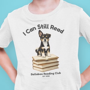 May include: White t-shirt featuring a black, brown, and white puppy sitting on a stack of books. The text "I Can Still Read" is above the puppy, and "Bellaboo Reading Club EST 2026" is below. The shirt is worn by a person.