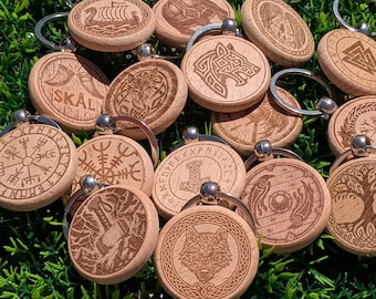 May include: A collection of round, light-colored wooden keychains featuring intricate laser-engraved designs. The engravings include various Norse-inspired symbols, such as the Vegvisir, Mjolnir, wolf heads, trees, and the word SKÅL, each attached to a metal ring.