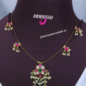 May include: A gold-toned necklace with a central pendant featuring a floral design with pink and green gemstones, and dangling green beads. The necklace has small pink gemstone accents and white pearl-like beads. The text "SBR10550" is visible.