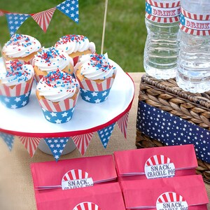 Pode incluir: Uma cena de piquenique patri&oacute;tica com cupcakes cobertos com glac&ecirc; branco e granulado vermelho, branco e azul. Os cupcakes est&atilde;o em um suporte branco para bolos, com sacos de lanches vermelhos rotulados "SNACK. CRACKLE. POP" abaixo. Garrafas de &aacute;gua com r&oacute;tulos "DRINK" e bandeirolas completam a cena.