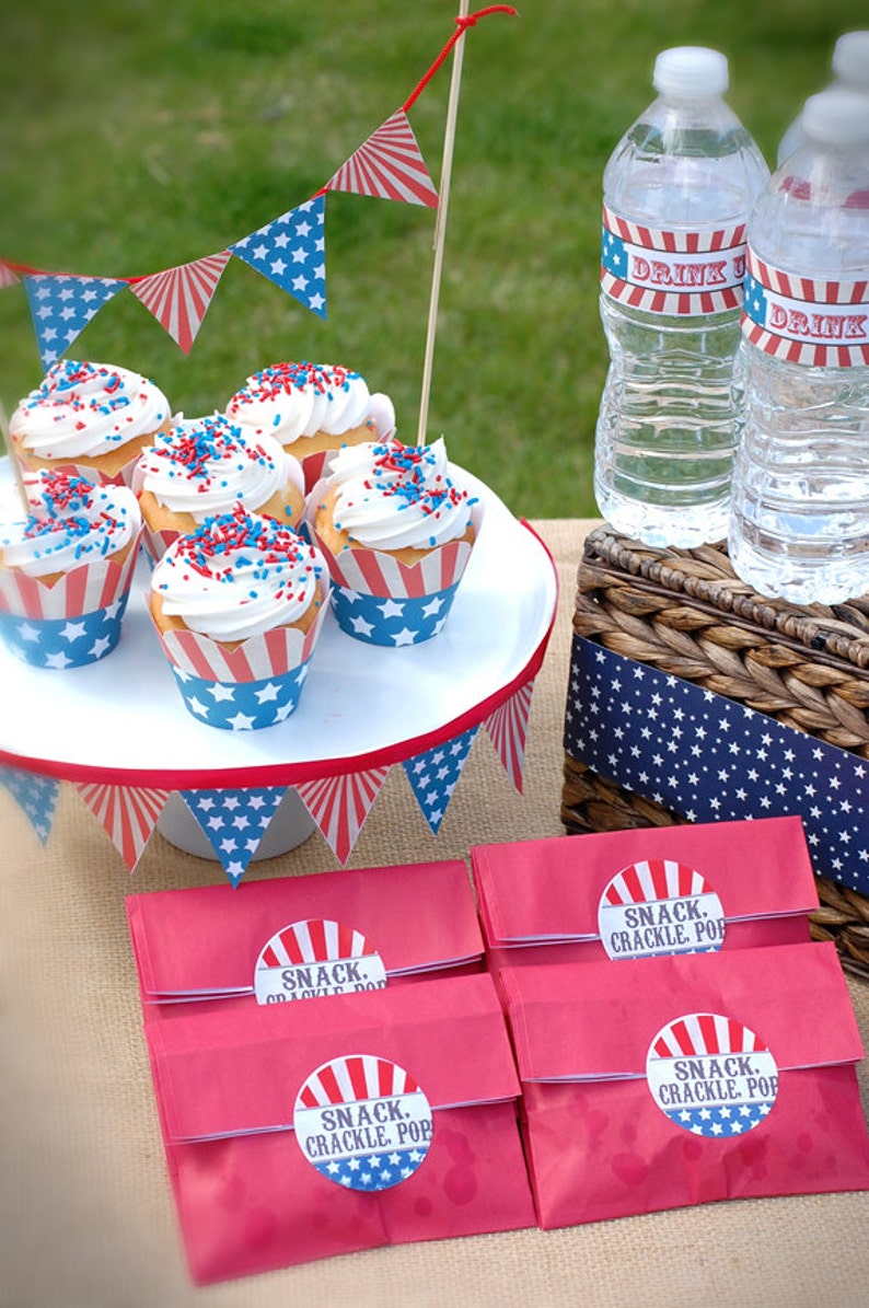 Pode incluir: Uma cena de piquenique patri&oacute;tica com cupcakes cobertos com glac&ecirc; branco e granulado vermelho, branco e azul. Os cupcakes est&atilde;o em um suporte branco para bolos, com sacos de lanches vermelhos rotulados "SNACK. CRACKLE. POP" abaixo. Garrafas de &aacute;gua com r&oacute;tulos "DRINK" e bandeirolas completam a cena.