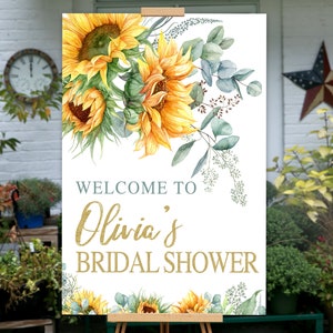 May include: Welcome to Olivia's Bridal Shower sign with a watercolor design of sunflowers and greenery. The text is in gold glitter.