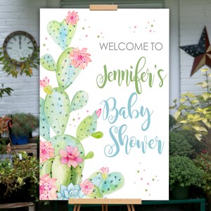 May include: A baby shower welcome sign featuring watercolor cacti with pink flowers and the text "Welcome to Jennifer's Baby Shower" in green and blue script. The sign is displayed on a wooden easel.