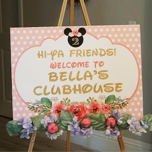 May include: A decorative sign with the text "Hi-Ya Friends! Welcome to Bella's Clubhouse" in gold lettering. The sign features a pink polka dot background, floral accents, and a Mickey Mouse silhouette. It is displayed on a wooden easel.