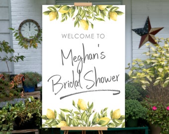 May include: A white sign on an easel with the text "WELCOME TO Meghan's Bridal Shower" in gray script. The sign is decorated with watercolor lemons and green leaves. The sign is outdoors, surrounded by plants and a clock.