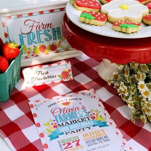 Puede incluir: Una exhibici&oacute;n con tem&aacute;tica de mercado de agricultores con fresas frescas, galletas y un letrero que dice "Farm Fresh". Un mantel a cuadros rojos y blancos prepara el escenario para una invitaci&oacute;n de cumplea&ntilde;os. Las galletas est&aacute;n decoradas para parecer fresas.