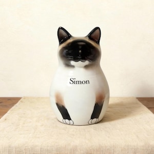 Op de afbeelding: Een keramieken Siamese kattenbeeldje met de naam "Simon" op de voorkant. De kat heeft een wit lichaam met bruine en zwarte aftekeningen op het gezicht, de oren en de poten. Het beeldje staat op een beige tafelloper.