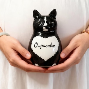 Pode incluir: Estatueta de gato de cerâmica preta e branca com a palavra "Chupacabm" no peito. O gato tem um desenho em forma de coração branco e é segurado em frente a uma pessoa vestindo um vestido branco.
