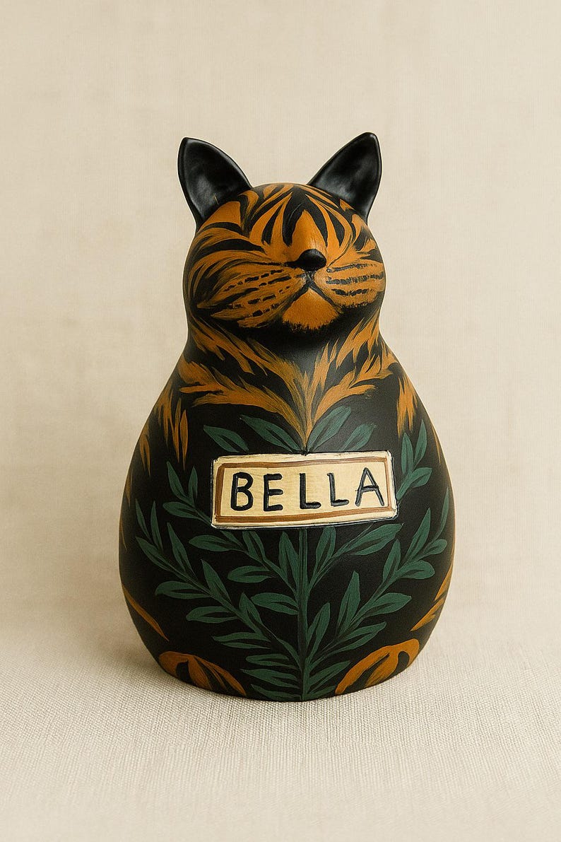 May include: A ceramic cat figurine with a tiger-striped head and black ears. The body is black with green foliage and a rectangular sign that reads "BELLA".