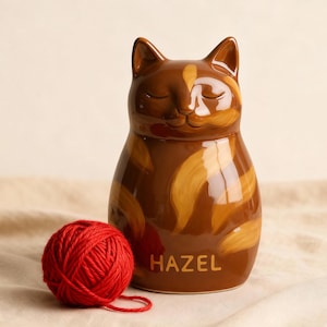 Puede incluir: Una figura de gato de cerámica marrón con una madeja de lana roja. El gato tiene los ojos cerrados y un patrón a rayas en tonos marrones y beige. El nombre "HAZEL" está impreso en la parte delantera.